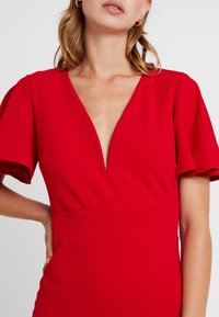 Czerwona sukienka z krótkimi bufkami, dekoltem w serek i dopasowaną talią. Gładki materiał z minimalną ilością szw�ów, eksponujący smukłą sylwetkę.