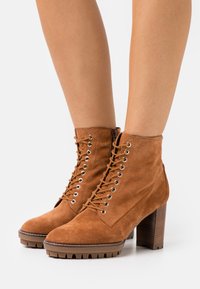 Stivaletti in suede marrone alla caviglia con lacci sul davanti, occhielli in metallo, tacco in legno spesso e suola in gomma texturizzata.