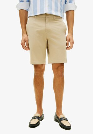 Homme portant un short beige mi-genou, une chemise rayée bleu clair et blanc avec les manches retroussées, et des mocassins noirs et beiges.