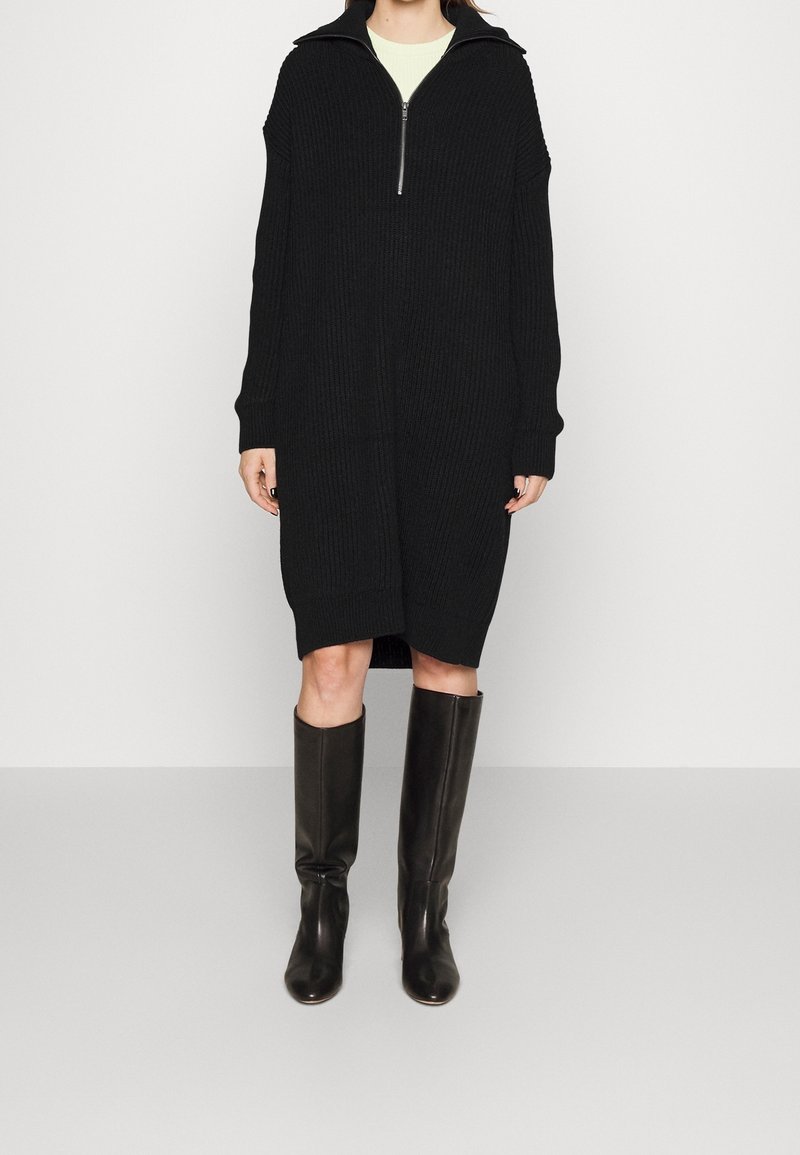 Robe oversize en maille côtelée noire avec un col montant zippé, dotée de manches longues et de poches latérales. Associée à des bottes en cuir noir montantes.
