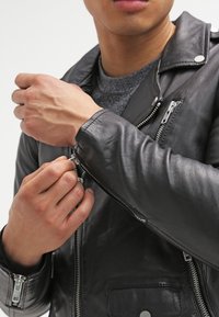 Svart lädermotorscykeljacka med silverdragkedjor, tryckknappar, fickor med dragkedja och en lätt strukturerad yta, bärs stängd vid kragen.