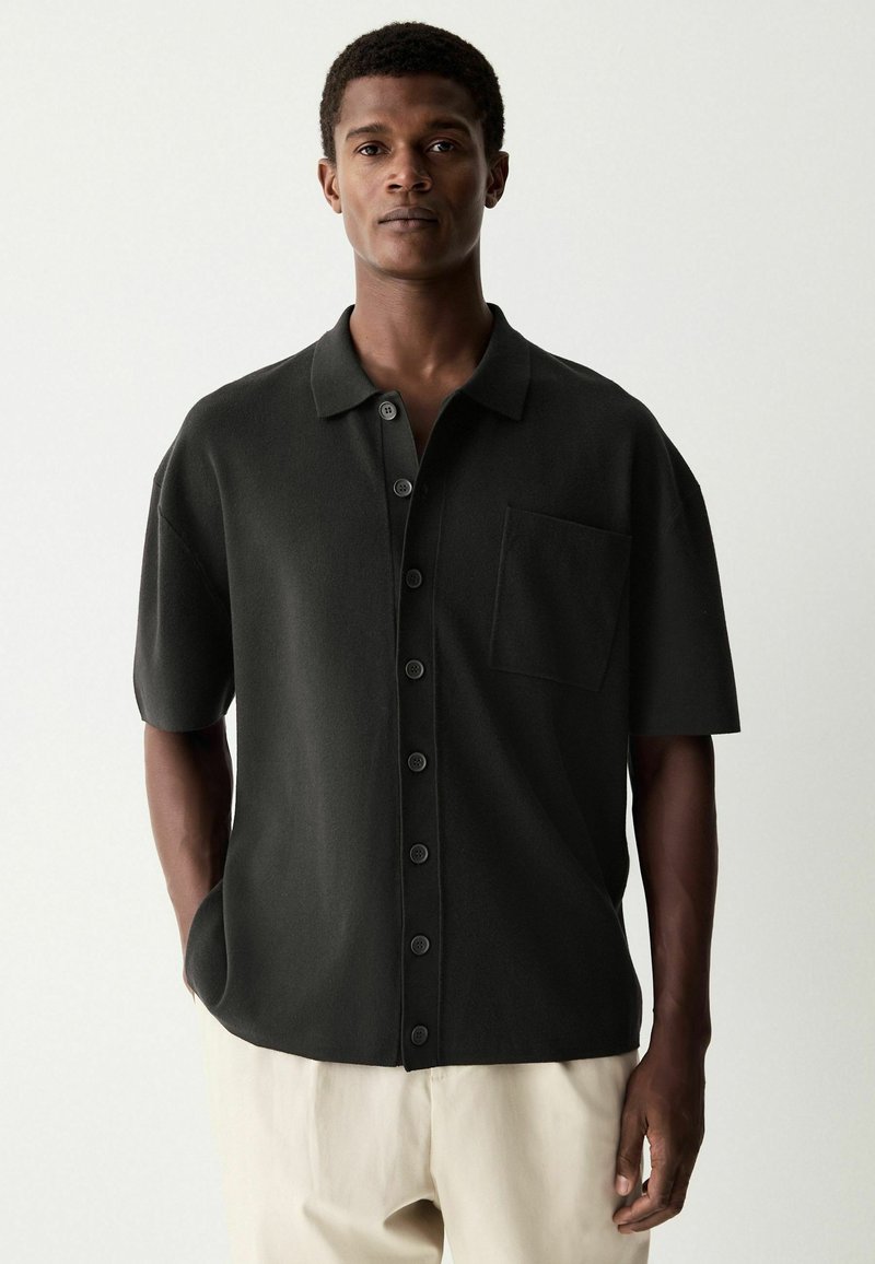 Camisa de manga corta negra en un tejido texturizado, con un cierre frontal de botones, un bolsillo y un diseño de cuello tradicional.