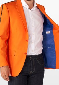 Jasnopomarańczowy żakiet o dopasowanym kroju, z dwoma guzikami i czarnymi paskami akcentującymi. Wnętrze ma niebieską podszewkę i małą kieszonkę.
