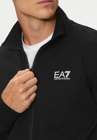 Giacca nera con zip, dotata di collo alto, texture morbida e logo bianco EA7 Emporio Armani sul petto.