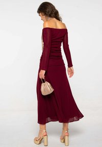 Robe bordeaux à décolleté bardot avec texture froncée, jupe fluide et petit sac beige. Comprend un collier en argent et des talons à lanières.