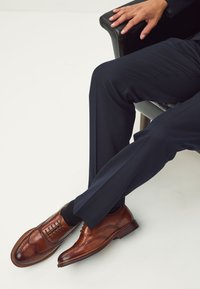 Chaussures habillées en cuir marron avec un détail brogue, associées à un pantalon sombre taillé sur mesure, montrant une texture lisse et un design classique.