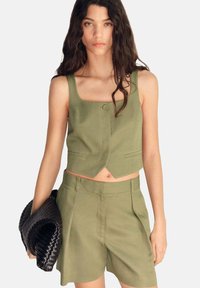 Olivově zelený cropped top s čtvercovým výstřihem a zapínáním na knoflíky, v kombinaci s odpovídajícími šortkami s vysokým pasem. Černá pletená kabelka.