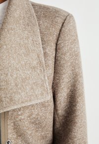 Beige teksturoitu takki, jossa on suuri kaulus, pehmeä kangas ja vetoketjullinen edessä. Mallissa on hillittyjä värisävyvaihteluita kauttaaltaan.