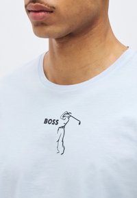 Κοντινό πλάνο ενός ατόμου που φοράει λευκό πουκάμισο με μια μινιμαλιστική μαύρη γραμμή σχεδίου ενός γκολφέρη που κάνει κούνια και τη λέξη «BOSS» τυπωμένη δίπλα του.