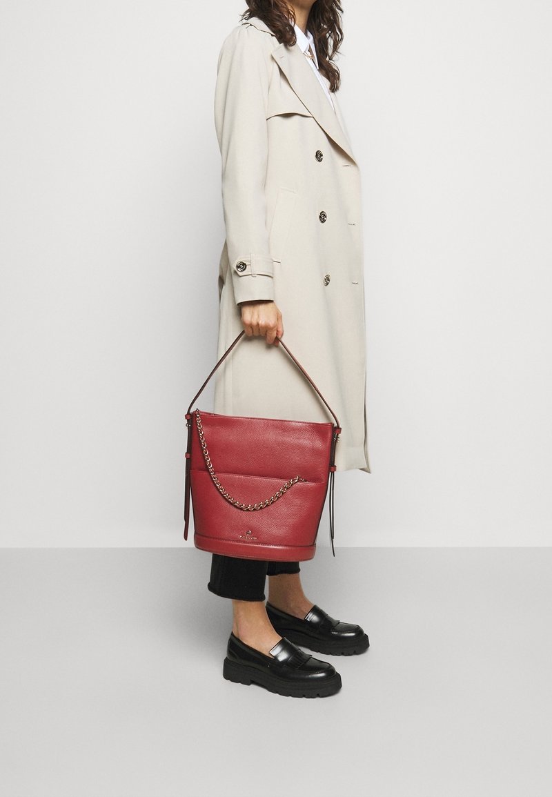 Femme portant un trench-coat beige et des mocassins noirs tenant un grand sac seau en cuir rouge avec un détail chaîne.