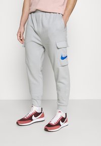 Svetlošedé cargo tepláky s elastickým pásom, bočnými kapsami a modrým logom Nike. Skombinované s červenými a bielymi športovými topánkami Nike.