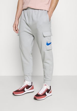 Pantaloni cargo grigi chiari in felpa con vita elastica, tasche laterali e logo Nike blu. Abbinati a scarpe da ginnastica Nike rosse e bianche.