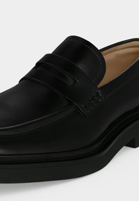 Mocassin en cuir noir avec une bride penny, bout arrondi et talon bas, montrant des coutures détaillées et une doublure intérieure couleur fauve.