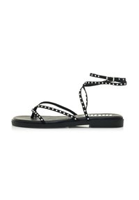 Sandalias planas negras con múltiples tiras que presentan pequeños tachuelas plateadas, un diseño de entrepiro y una correa ajustable en el tobillo. Acabado mate suave.