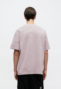 Ανοιχτό ροζ κοντομάνικη μπλούζα, ύφασμα από μείγμα βαμβακιού, χαλαρή εφαρμογή, στρογγυλή λαιμόκοψη, ορατές ραφές, απλή πλάτη χωρίς διακοσμητικά στοιχεία.