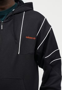 Μαύρο φούτερ με κουκούλα Adidas με λευκές γραμμές, πορτοκαλί λογότυπο στο στήθος, φερμουάρ μπροστά και κουκούλα με κορδόνια σε άτομο που φοράει σκούρο πράσινο πουκάμισο από κάτω.