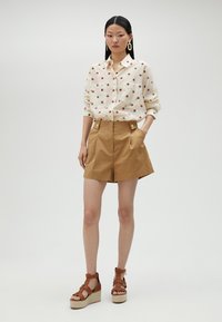 White button-up shirt with red polka dots, tan high-waisted shorts with pockets, and brown strappy wedge sandals.