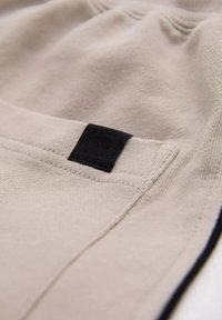 Tissu beige avec une texture lisse et une petite étiquette noire sur la poche. Présente des coutures contrastées le long des bords et une coupe décontractée.