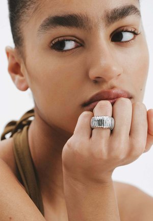 Anillo de plata con textura estriada, llevado en una mano, mostrando un acabado brillante. Tono de piel visible en la mano y la cara, con un fondo neutral.