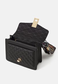 Sac à main matelassé noir avec un fermoir en or, doté d'une doublure intérieure à motifs et d'un foulard décoratif qui dépasse. Design compact.