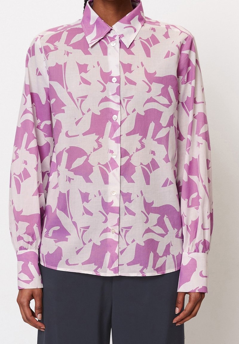 Chemise à manches longues boutonnée avec un motif abstrait violet et blanc, portée avec un pantalon sombre.