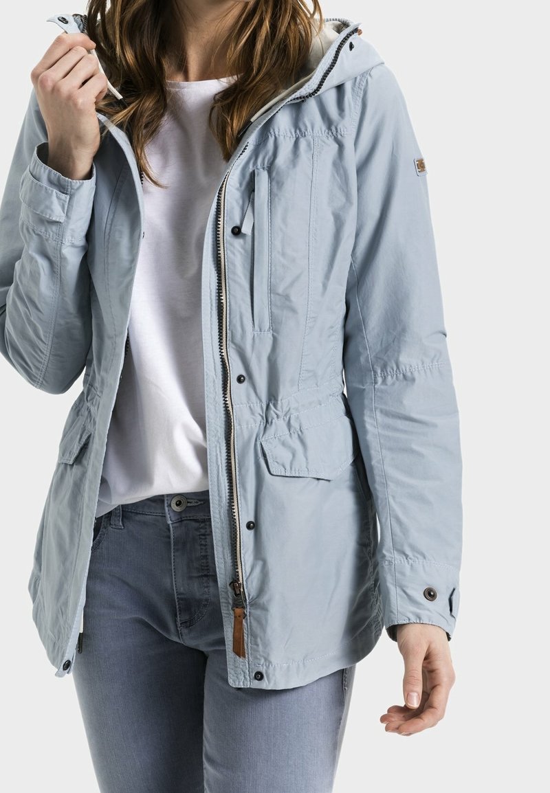 Femme portant une veste à capuche bleu clair, une chemise blanche et un jean bleu, tenant le cordon de la veste près du col.