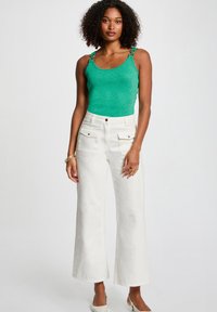 Groen metallic tanktop met decoratieve bandjes, gecombineerd met witte wijde broek met voorzakken en details in goudkleurig metaal.