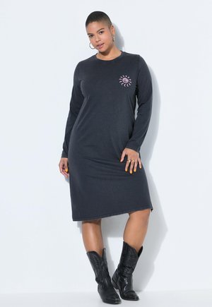 Donna in piedi contro un muro che indossa un vestito nero a maniche lunghe con un motivo di sole rosa e stivali da cowboy neri, con smalto giallo visibile.