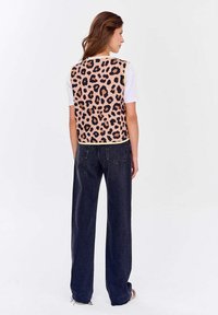 Žena s dlouhými hnědými vlasy má na sobě růžovou vestu s leopardím potiskem přes bílé triko s krátkým rukávem a volné černé džíny, stojí proti bílému pozadí.