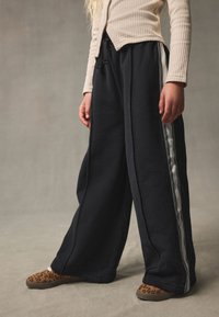 Schwarze Sweatpants mit weitem Bein und grauem Seitenstreifen, ausgestattet mit elastischem Bund und Kordelzug. Kombiniert mit Leopardenmuster-Slippern.