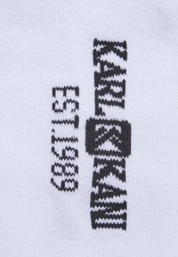 Biela pletená látka s čiernym vyšívaným textom a vzormi, vrátane stilizovaného dizajnu a loga. Textúra sa zdá byť hladká a konzistentná.