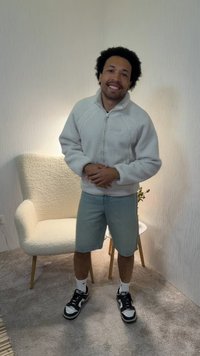 Άνδρας που στέκεται σε εσωτερικό χώρο, φοράει ελαφρύ φλις μπουφάν, σορτς, αθλητικά παπούτσια, χαμογελάει, με υφασμάτινη πολυθρόνα, μικρό τραπέζι και φυτό στο παρασκήνιο.