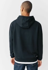 Schwarzer Kapuzenpullover aus weichem Stoff, mit gerippten Bündchen und Saum. Sichtbare Steppnähte, getragen über einem weißen T-Shirt und Jeans.