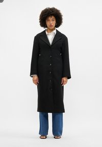 Dlouhý černý kabát s klopovým límcem, zapínáním na knoflíky a dvěma předními kapsami. Nositý přes bílou košili a modré široké džíny.