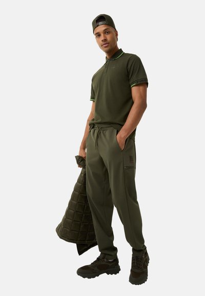 Olive green polo shirt with contrasting collar, matching joggers with side zip pocket, and insulated jacket in quilted texture. Dark footwear.