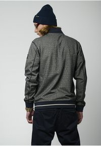 Giacca grigia con motivo, colletto e polsini a coste blu navy, abbinata a un berretto blu navy. Presenta una vestibilità slim e un dettaglio contrastante in vita.