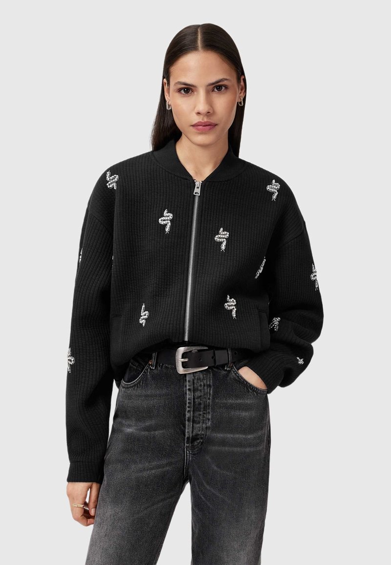 Sort zip-jakke med ribbenstruktur og slangebroderi. Sammen med højtaljede mørkegrå jeans med en metalbæltespænde.