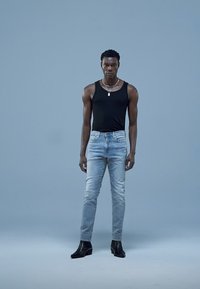 Mustast tanktopist, helesinist kulunud teksadest, kitsas lõige; modell kannab musti kontsaga saapaid; neutraalne taust, suunatud valgustus.