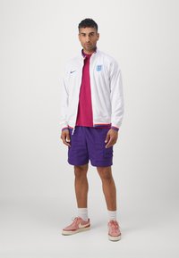 Biela zipsová bunda s červenými a modrými akcentmi, vínové tričko, fialové šortky a ružové tenisky. Obsahuje logo Nike a erb Anglicka.