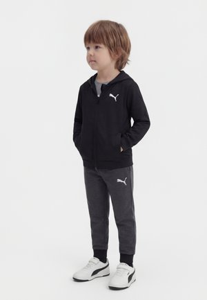 Niño pequeño de pie mirando hacia la izquierda, vestido con sudadera con capucha negra con cremallera, pantalones deportivos gris oscuro y zapatillas blancas con detalles negros.