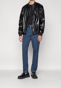 Veste bomber en simili cuir noir avec un motif en relief, associée à un pantalon chino bleu et des chaussures noires épaisses, avec une ceinture simple.