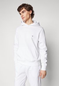 Haut à capuche blanc en tissu mélangé coton, avec une poche kangourou à l'avant et un petit logo sur la poitrine. Texture lisse, coupe décontractée.