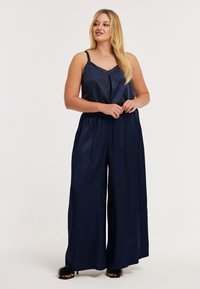 Marineblauwe satijnen jumpsuit met een V-hals, dunne bandjes, elastische tailleband en wijde pijpen. Gestructureerde stof voegt een subtiele elegantie toe.