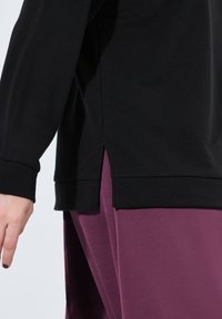 Svart långärmad topp med en sidoflik och ribbade ärmslut, kombinerad med mjuka, lila byxor. Tyget verkar slätt och elastiskt.