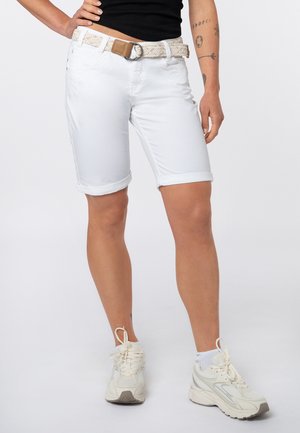 Persoon draagt witte knielange shorts met een gevlochten riem, zwarte top, witte sneakers en zichtbare armtatoeages op een effen achtergrond.