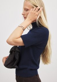 Tumes ribakudumine sveater kõrge kaeluse ja lühikeste tekstureeritud varrukatega, koos musta käekotiga ja hõbedase lainelise käevõruga.