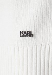 Baltas megztinis, turintis raukšlėtą tekstūrą ir juodą logotipo pin'ą, kuriame parašyta "KARL LAGERFELD", kairėje pusėje.