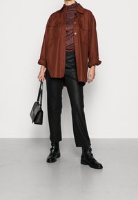 Chemise oversize marron avec de grandes poches poitrine, associée à un haut à motif et un pantalon en simili cuir noir. Complétée par des bottines noires et un sac.