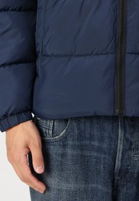 Navy blue puffer jacket with a zippered front and elasticized cuffs, paired with light blue denim jeans. Smooth texture, quilted design.