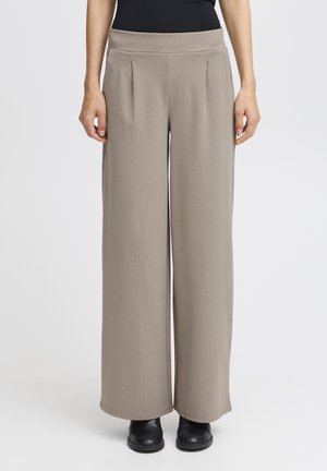 Helle braune weit geschnittene Hose mit Falten und einem flachen Bund, aus einem weichen, elastischen Stoff; kombiniert mit schwarzen Knöchelstiefeln.